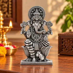 Front view of antique silver Lord Ganesha murti on a wooden pooja table with a brass diya, flowers, ornate crown and lotus base.